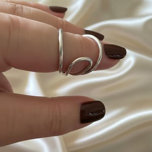 Puede incluir: Un anillo plateado con un dise&ntilde;o &uacute;nico y fluido. El anillo se envuelve alrededor del dedo con una forma curva y abierta. El anillo se muestra sobre un fondo suave y blanquecino.