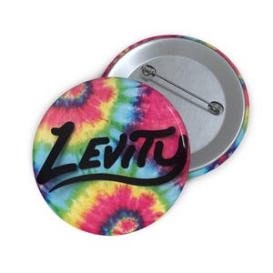 Puede incluir: Un botón redondo con un diseño tie-dye en tonos rosa, amarillo, azul y rojo. La palabra "Levity" está impresa en negro en cursiva en la parte delantera del botón. Se ve un segundo botón, que muestra la parte trasera del alfiler.