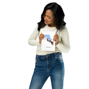 Puede incluir: Una mujer sostiene una tarjeta blanca con un recorte del país de Armenia. El recorte muestra una cordillera y una iglesia. El texto "Armenia" está escrito debajo del recorte.