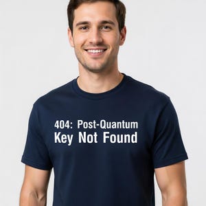 Peut inclure: Un t-shirt bleu marine avec le texte "404: Post-Quantum Key Not Found" en blanc. Le t-shirt est porté par une personne souriante.