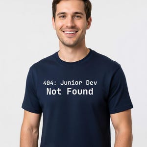 Peut inclure: T-shirt bleu marine avec l'inscription blanche "404: Junior Dev Not Found". Le t-shirt est à col rond et est porté par une personne.