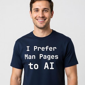 May include: Navy blue t-shirt with the white text "I Prefer Man Pages to AI." The shirt is short-sleeved and has a crew neck. The person wearing the shirt is smiling.