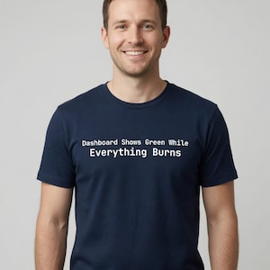 May include: A navy blue short-sleeved t-shirt featuring white text printed on the chest that reads: Dashboard Shows Green While Everything Burns. The shirt has a crew neckline and a standard fit, providing a clean and simple appearance.