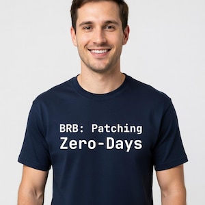 May include: Navy blue t-shirt with the white text "BRB: Patching Zero-Days". The shirt is a short-sleeved crew neck style. The person in the image is smiling.