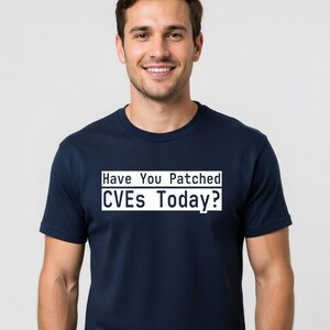 Op de afbeelding: Marineblauwe t-shirt met de tekst "Have You Patched CVEs Today?" in een witte rechthoek. Het t-shirt heeft een ronde hals en wordt door een persoon gedragen.