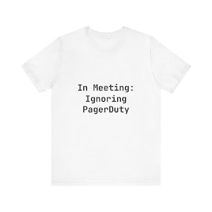 Könnte beinhalten: Weißes T-Shirt mit dem Text "In Meeting: Ignoring PagerDuty" in Schwarz, zentriert auf der Vorderseite. Das Shirt hat kurze Ärmel und einen Rundhalsausschnitt.