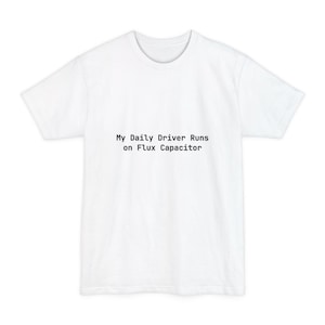 以下が含まれることがあります： 「My Daily Driver Runs on Flux Capacitor」の黒い文字がプリントされた白いTシャツ。クルーネックと半袖のデザインです。テキストはシャツの前面中央に配置されています。
