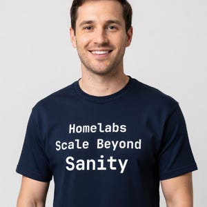 Puede incluir: Una camiseta azul marino con el texto blanco "Homelabs Scale Beyond Sanity". La camiseta la lleva una persona sonriendo a la cámara. El fondo es blanco liso.
