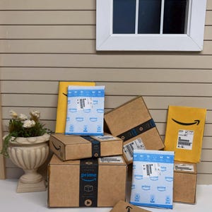 May include: A collection of miniature Amazon Prime packages and envelopes. The packages are brown cardboard boxes with black tape and the Prime logo. The envelopes are yellow and blue with the Amazon logo and shipping labels.
