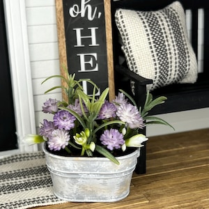 May include: A miniature galvanized metal bucket filled with purple and white flowers and green foliage. The bucket sits on a wooden surface, with a black and white patterned pillow and a sign that says "oh, HE" in the background.