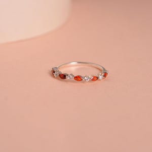 May include: A silver ring with a row of red and white gemstones. The ring is on a light pink background.