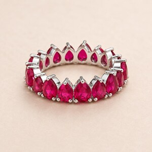 May include: A silver-toned eternity band featuring a continuous row of vibrant, pear-shaped red gemstones. Each stone is individually set in a prong setting, creating a circular design that showcases the faceted surfaces of the red gems.