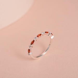May include: A silver ring with alternating red and clear gemstones. The red gemstones are marquise-cut, and the clear gemstones are round. The ring is set against a soft pink background.