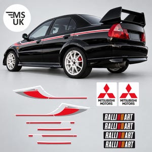 May include: A black saloon car with a rear spoiler and white multi-spoke wheels, featuring red and white side body decals. Below the car are additional decal sets including two Mitsubishi Motors logos and four Ralliart text decals with red and orange stripes.