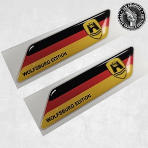 以下が含まれることがあります： 「WOLFSBURG EDITION」の文字が入った、黒、赤、黄色の光沢のあるステッカー2枚。各ステッカーには、ドイツ国旗を連想させるシールドエンブレムとデザインが施されています。ステッカーは白い台紙に貼られています。