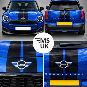 Op de afbeelding: Een blauwe Mini Countryman met zwarte racestrepen en een dunne blauwe middenlijn over de motorkap en achterklep. De auto is voorzien van het Mini-logo aan de voor- en achterzijde, met de tekst COUNTRYMAN op de achterklep.