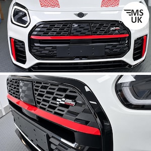 Op de afbeelding: De voorzijde van een witte Mini Cooper met een zwarte honingraatgrille, een horizontale rode accentbalk, een John Cooper Works-embleem, rood geblokte stickers op de motorkap en een zwarte houder voor de kentekenplaat.