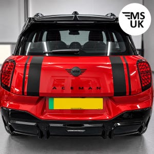 Op de afbeelding: Een felrode Mini Aceman hatchback gezien vanaf de achterkant, met een zwart dak, zwarte achterspoiler en twee verticale zwarte racestrepen. Het voertuig heeft een zwart Mini-embleem, zwarte belettering en een geel kentekenplaat.