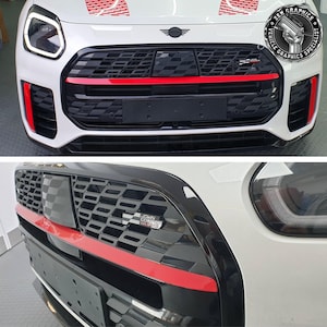 Op de afbeelding: Close-up van de grille van een witte auto met rode accenten. De grille heeft een zwart honingraatpatroon met een rode horizontale streep. De auto heeft een zwart kentekenplaatgedeelte en de tekst "JOHN COOPER WORKS".