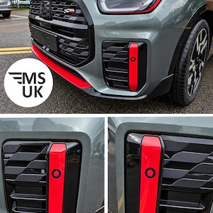 May include: A sage green vehicle front bumper featuring a black grille with a small checkered emblem, bright red vertical accent strips on the air intakes, and a red lower lip spoiler. The design includes integrated circular sensors.