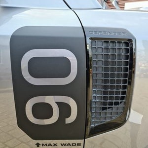 以下が含まれることがあります： 黒いグリルと黒いステッカーが付いた灰色の車。ステッカーには「DEFENDER」と「90」の文字が書かれており、上向きの矢印と「MAX WADE」の文字が下に書かれています。