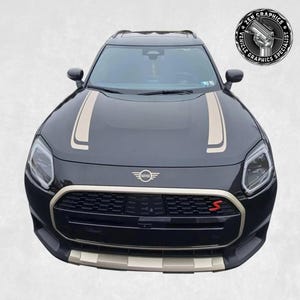 Może przedstawiać: Czarny Mini Cooper Countryman z złotymi paskami na masce i czerwoną odznaką "S" na przednim grillu.