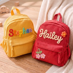 May include: Two corduroy backpacks, one yellow and one red. The yellow backpack features the name Stephen in multi-colored yarn letters. The red backpack features the name Hailey in cream yarn letters with a small flower and a cherry applique.