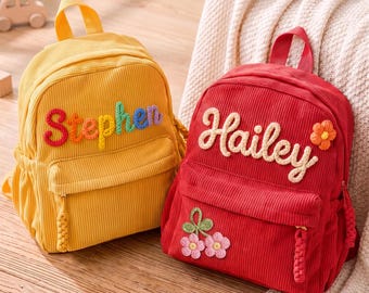 May include: Two corduroy backpacks, one yellow and one red. The yellow backpack features the name Stephen in multi-colored yarn letters. The red backpack features the name Hailey in cream yarn letters with a small flower and a cherry applique.