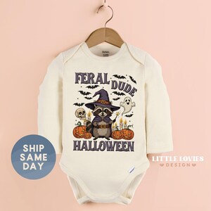 May include: Cream-colored baby bodysuit with long sleeves. The front features a Halloween-themed graphic with a raccoon wearing a witch hat, surrounded by pumpkins, bats, and the text "FERAL DUDE HALLOWEEN".