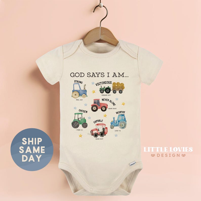 A baby onesie with a design of various farm vehicles, including tractors, trucks, and plows, along with the text "God says I am..." The onesie is hanging on a wooden hanger against a pink background.
