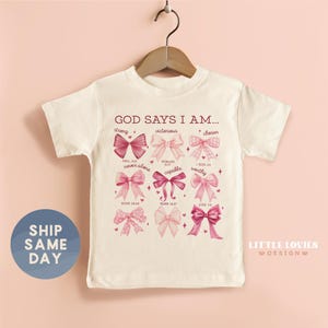 May include: A white t-shirt with the text "GOD SAYS I AM..." and various bow designs in shades of pink. The shirt includes phrases like "strong," "victorious," and "chosen." The shirt is hanging on a wooden hanger.