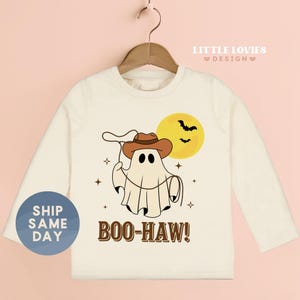 May include: Cream-colored long-sleeve shirt featuring a cartoon ghost wearing a cowboy hat and holding a lasso. The design includes a yellow moon, bats, and the text "BOO-HAW!" A blue circle on the left says "SHIP SAME DAY".