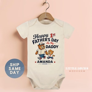 May include: Cream-colored baby bodysuit with the text "Happy 1st Father's Day As My Daddy" and a cartoon bear illustration. The design includes the name "AMANDA" and the year "2026". The bodysuit is hanging on a wooden hanger.