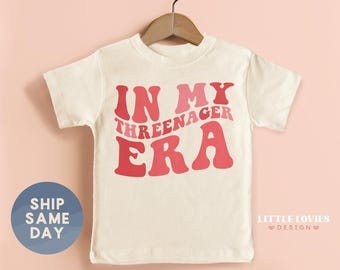 May include: Off-white toddler t-shirt with the text "IN MY THREENAGER ERA" in shades of red. The shirt is hanging on a wooden hanger against a pink background. A blue circle with the words "SHIP SAME DAY" is on the left.
