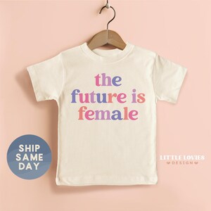 A white t-shirt with the text "the future is female" printed on it, hanging on a wooden hanger against a peach background.