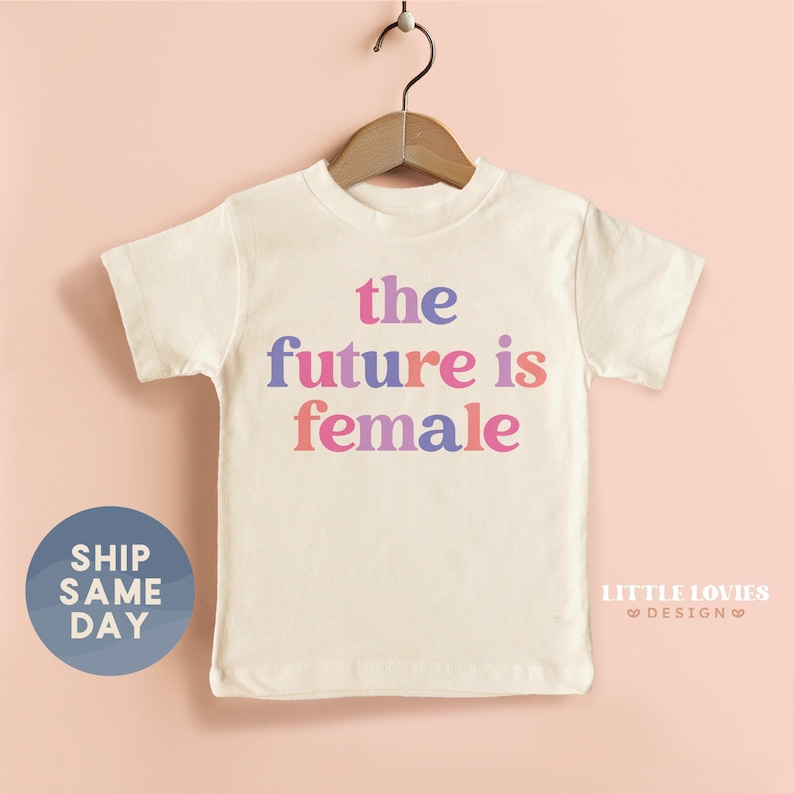 A white t-shirt with the text "the future is female" printed on it, hanging on a wooden hanger against a peach background.