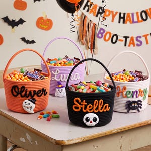 Könnte beinhalten: Vier runde Seilkörbe für Halloween-Süßigkeiten, jeder in einer anderen Farbe und mit personalisiertem Namen. Der orangefarbene Korb trägt den Namen Oliver, der lila Korb Luna, der schwarze Stella und der weiße Henry, mit festlichen Applikationen.