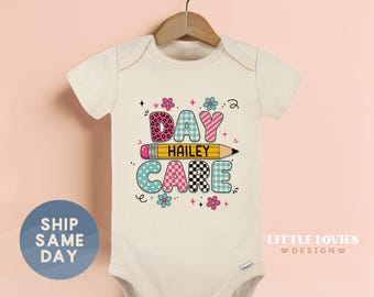 May include: A cream-colored short-sleeve baby bodysuit featuring a graphic print with the text DAY CARE and the name HAILEY centered on a yellow pencil illustration. The letters are decorated with various patterns including leopard print and plaid.