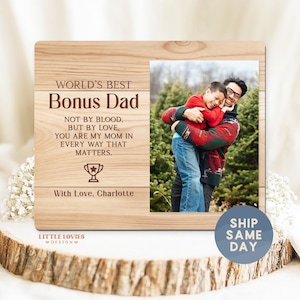 May include: Wooden picture frame with a photo of a man and a child hugging. The frame has the text "WORLD'S BEST Bonus Dad" and a message below. The frame is on a wooden surface.