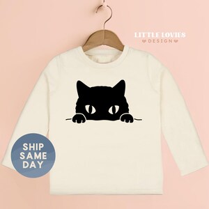 A white long-sleeved t-shirt with a black cat graphic printed on it, hanging on a wooden hanger.