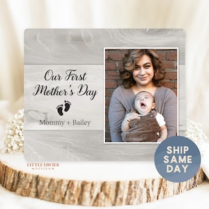 May include: A wooden photo frame with a light gray wood grain design. The left side has the text "Our First Mother's Day", footprints, and "Mommy + Bailey". The right side displays a photo of a mother and baby. The frame is on a wooden base.