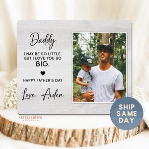 May include: A wooden picture frame with a photo of a father and child. The frame has the words "Daddy," "I may be so little, but I love you so BIG," "Happy Father's Day," and "Love, Aiden." The frame is on a wooden surface.