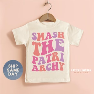 May include: A cream-colored t-shirt with the words "SMASH THE PATRIARCHY" in a retro, colorful font. The text is in shades of pink, purple, and orange. The shirt is hanging on a wooden hanger.
