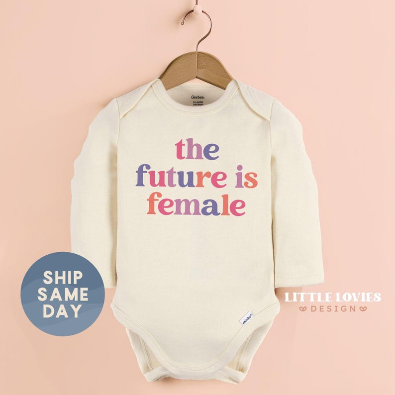 A white baby onesie with the text "the future is female" printed on it, hanging on a wooden hanger against a peach background.