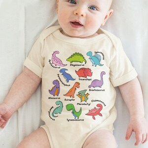 A baby wearing a white onesie with illustrations of various dinosaurs.