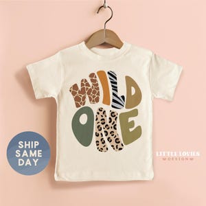 A white t-shirt with a graphic design featuring the text "WILD ONE" in a stylized font, surrounded by various animal prints such as leopard, zebra, and giraffe. The t-shirt is hanging on a wooden hanger against a pink background.