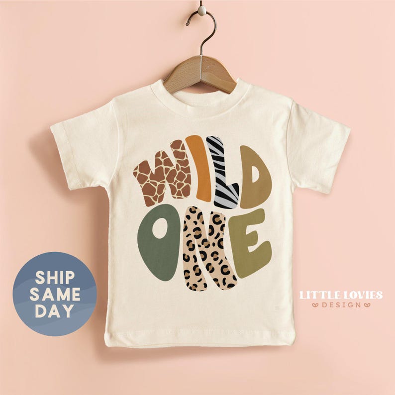 A white t-shirt with a graphic design featuring the text "WILD ONE" in a stylized font, surrounded by various animal prints such as leopard, zebra, and giraffe. The t-shirt is hanging on a wooden hanger against a pink background.