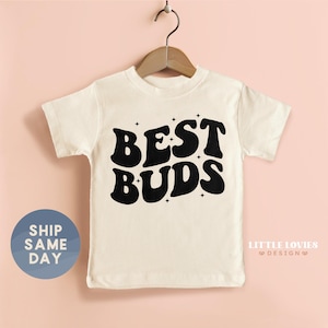 May include: Cream-colored t-shirt with the words "BEST BUDS" in bold, black, retro-style lettering. The shirt is hanging on a wooden hanger against a light pink background. The shirt has a round neck and short sleeves. A blue circle with the words "SHIP SAME DAY" is on the left.