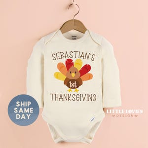 May include: Cream-colored baby bodysuit with long sleeves, featuring a Thanksgiving turkey graphic. The design includes the text "Sebastian's 1st Thanksgiving". The bodysuit is hanging on a wooden hanger.