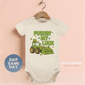 May include: Cream-colored baby bodysuit with a green tractor graphic and the text "Pushin' My Luck." The tractor has a leprechaun hat and is scooping up shamrocks. The bodysuit is hanging on a wooden hanger.